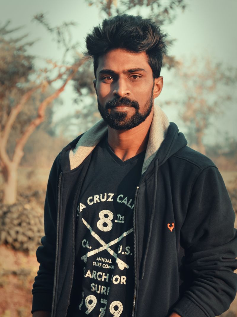 Representative portrait — A young South Asian man with a beard in a black hoodie poses confidently outdoors, surrounded by trees.