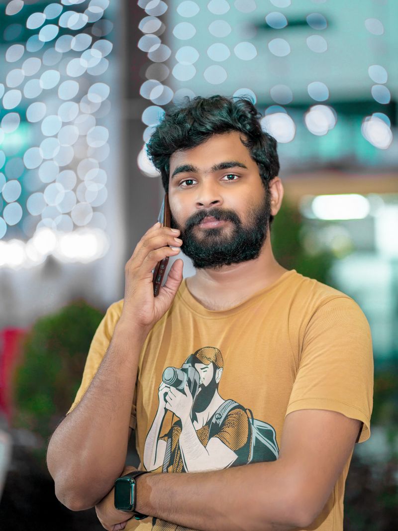 Representative portrait — Casual bearded man in a t-shirt making a call outdoors with festive lights in the background.