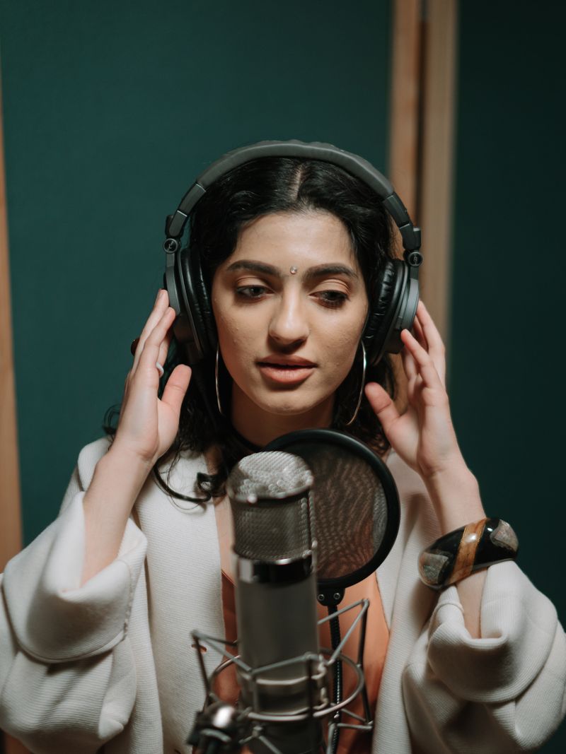 Representative portrait — A young woman sings into a microphone in a professional music studio setting, wearing headphones.